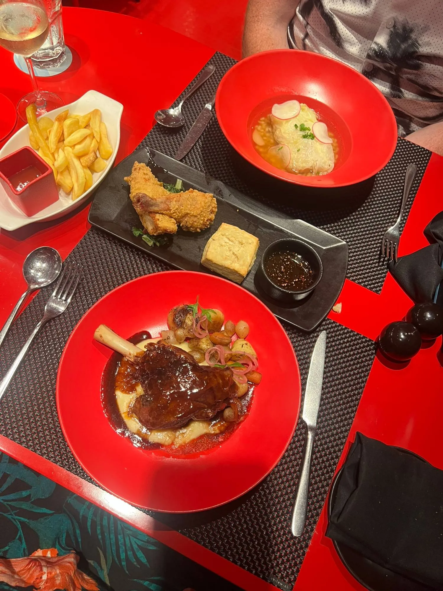 The mains on the red table: a lamb shank in rich gravy on mash in a red bowl, V fried chicken with an American biscuit and dipping sauce on a black slate, a bowl of chips, and the short rib pastrami in broth.