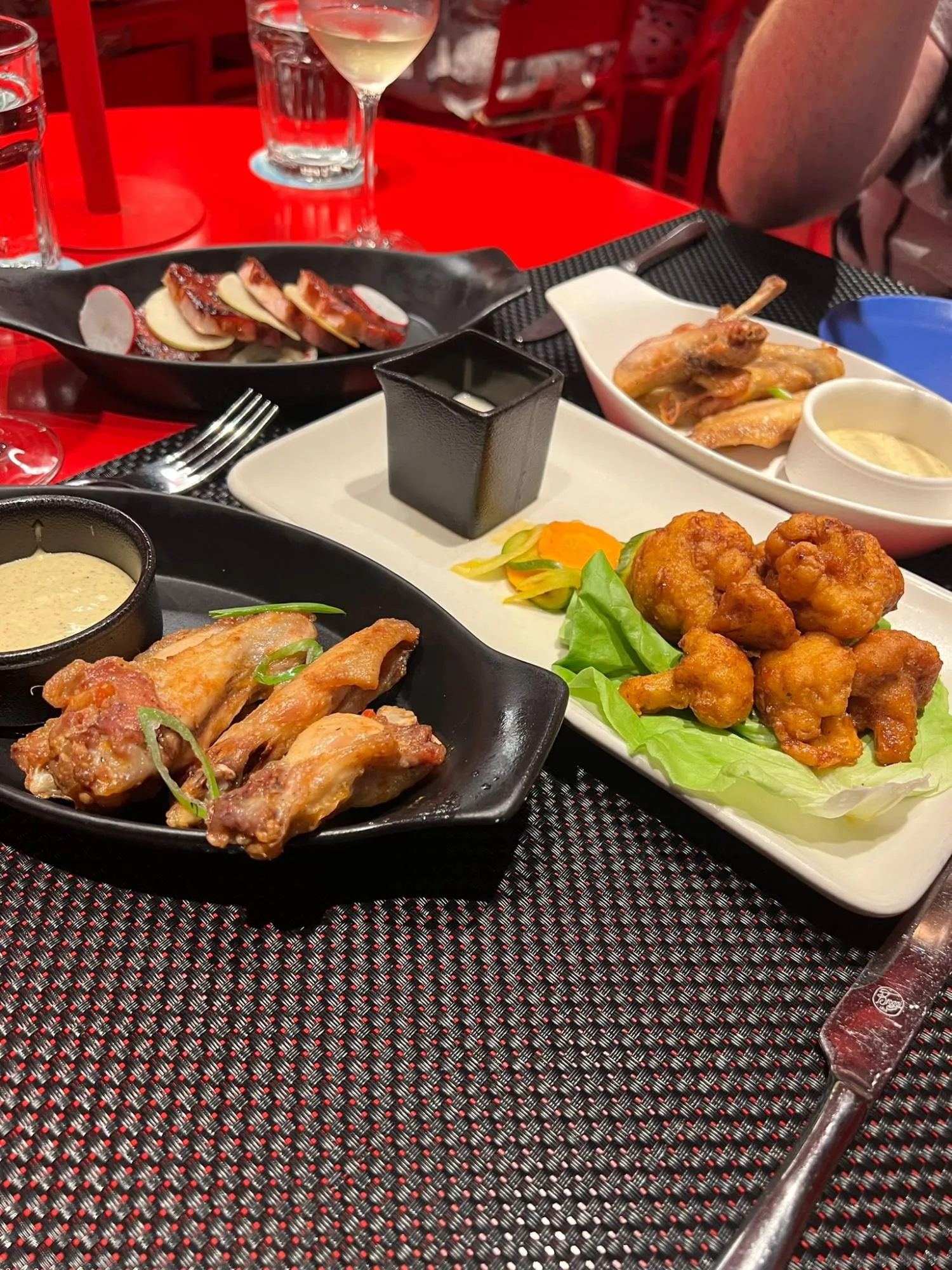 The starters at Razzle Dazzle laid out on a red table: chicken wings in a black dish with a creamy dip, maple glazed pork belly bites with apple slices, more wings with aioli, and Nashville hot cauliflower on lettuce.