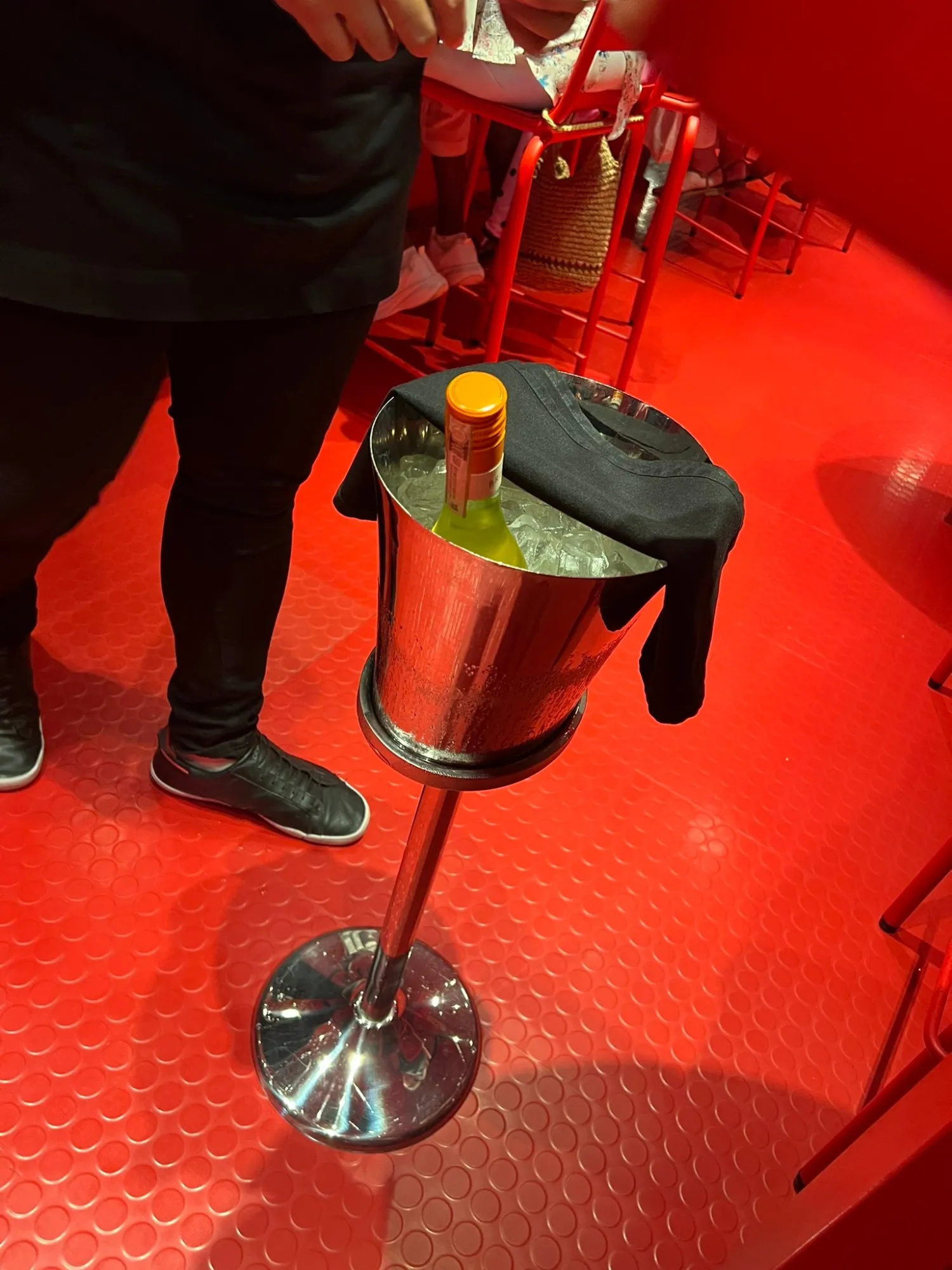 A bottle of white wine chilling in a silver ice bucket on a stand, against the bright red floor and red chairs of Razzle Dazzle.
