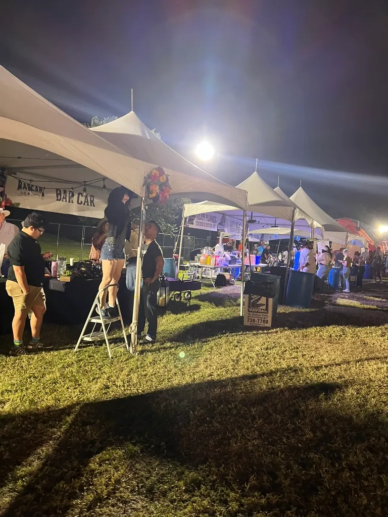 Rows of festival tents at night with people milling between them, someone up a step ladder decorating the Bar Car tent