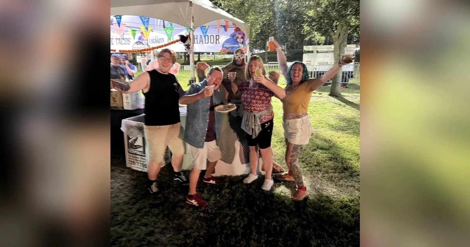 The four of us cheering with drinks in the air outside the Galaxie Tacos and El Luchador tent at night, papel picado bunting and a cardboard luchador cut-out behind us