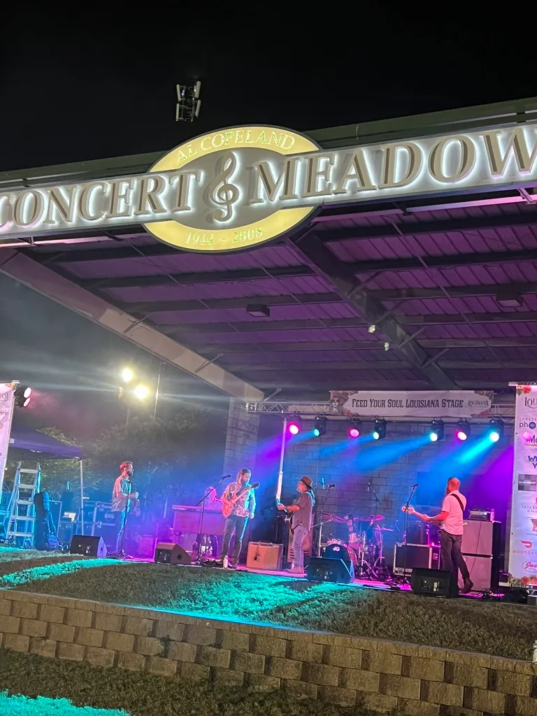 A five piece band on the Feed Your Soul Louisiana Stage under the Al Copeland Concert Meadow sign, lit with teal and blue stage lights