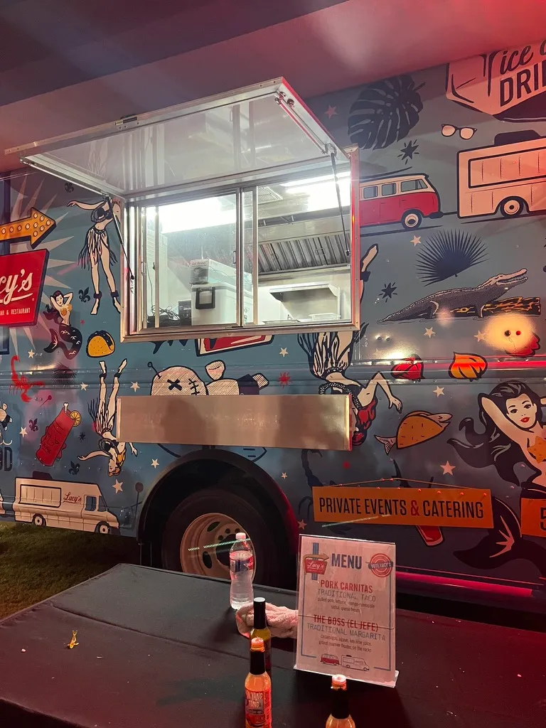 Lucy's food truck at night, a menu card on the table in front listing a pork carnitas traditional taco and The Boss el Jefe traditional margarita