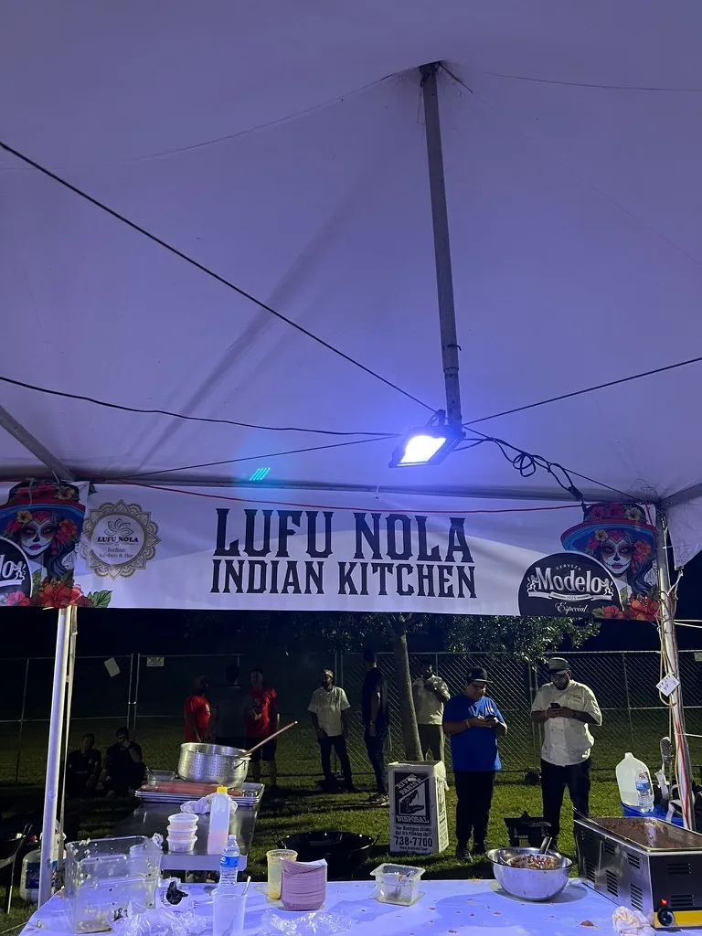 The Lufu Nola Indian Kitchen tent at night under a blue floodlight, staff standing behind a prep table with a large stockpot