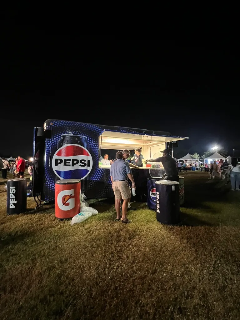 A Pepsi branded trailer at night with Gatorade and Pepsi barrels outside it and a few people at the serving window