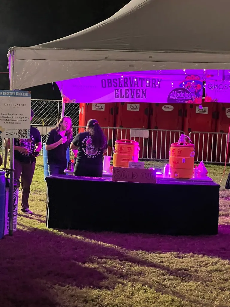 The Observatory Eleven tent under purple light, staff in branded t-shirts behind a black table with drinks dispensers and a handwritten SOLD OUT sign