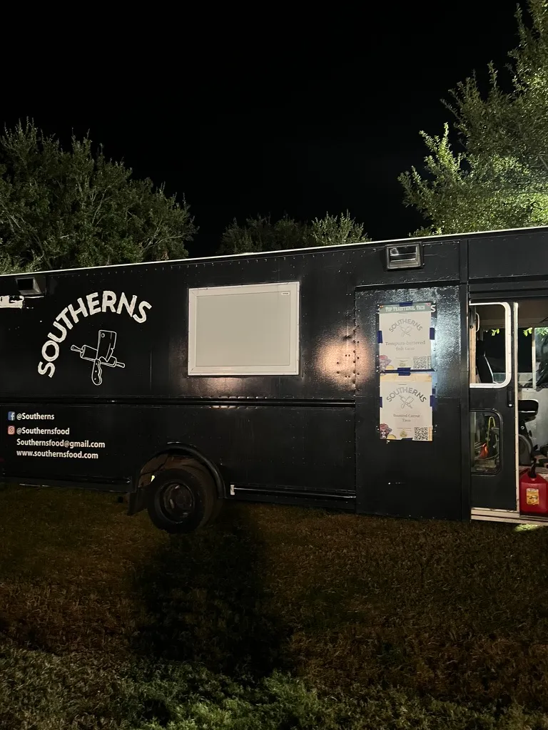 The black Southerns food truck at night with two printed posters taped to the side, one for a tempura battered fish taco and one for a roasted carrot taco, each with a QR code