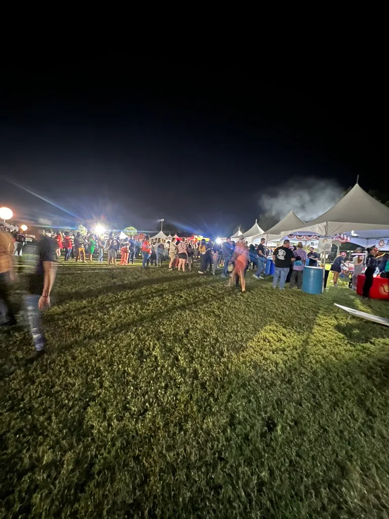 A row of white festival tents at night with a crowd of people between them, steam rising off a barbecue and floodlights flaring behind