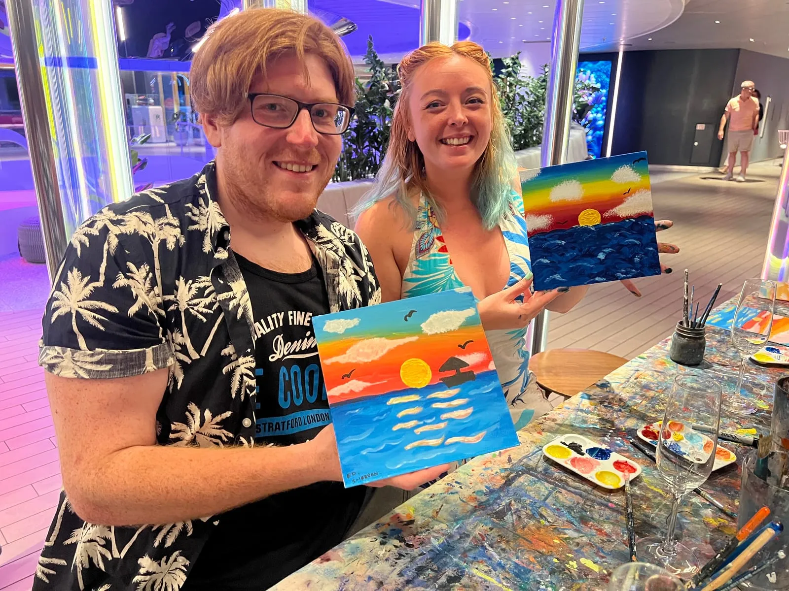 Us holding up our two finished paintings at the art class, both sunsets over the sea, done in the same session from the same instructions