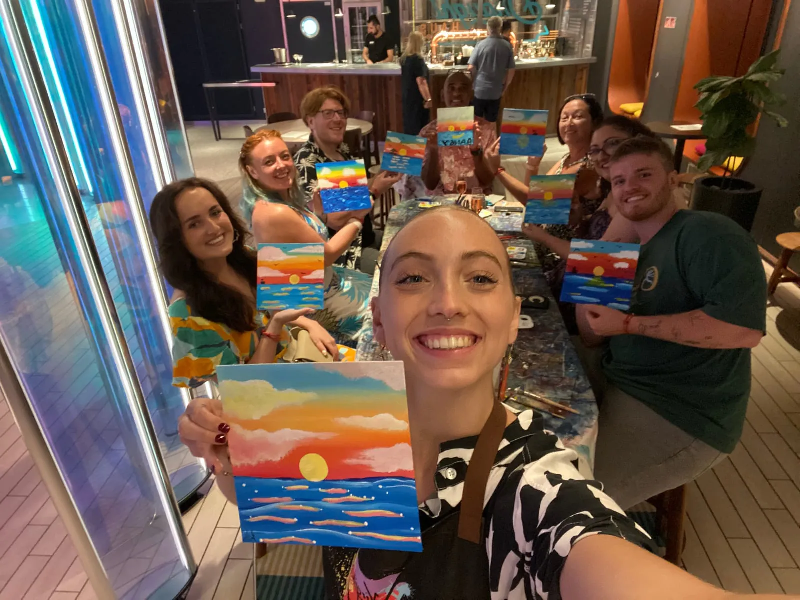 The whole class in Draught Haus holding up their finished paintings at the end of the session, every one of them the same sunset done differently