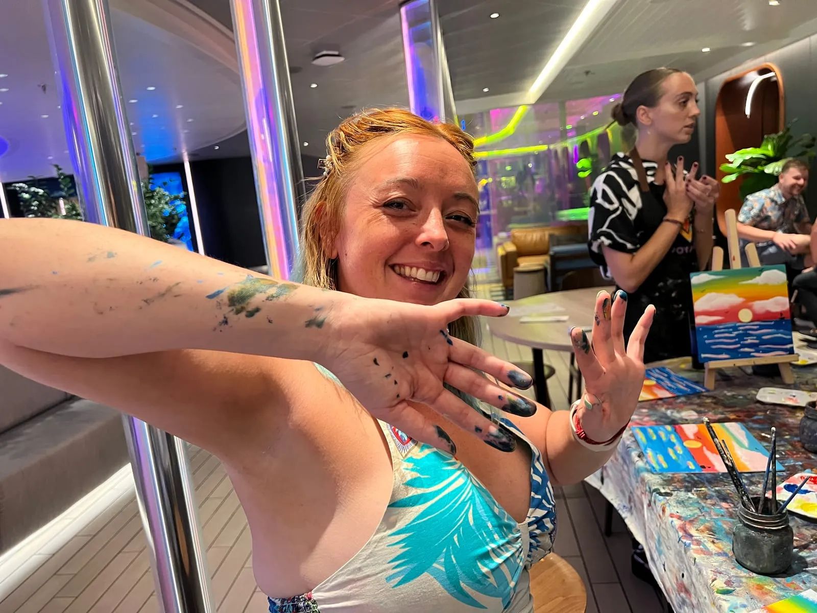 My hands and forearm covered in smudges of blue and green paint, with finished canvases drying on little wooden easels behind