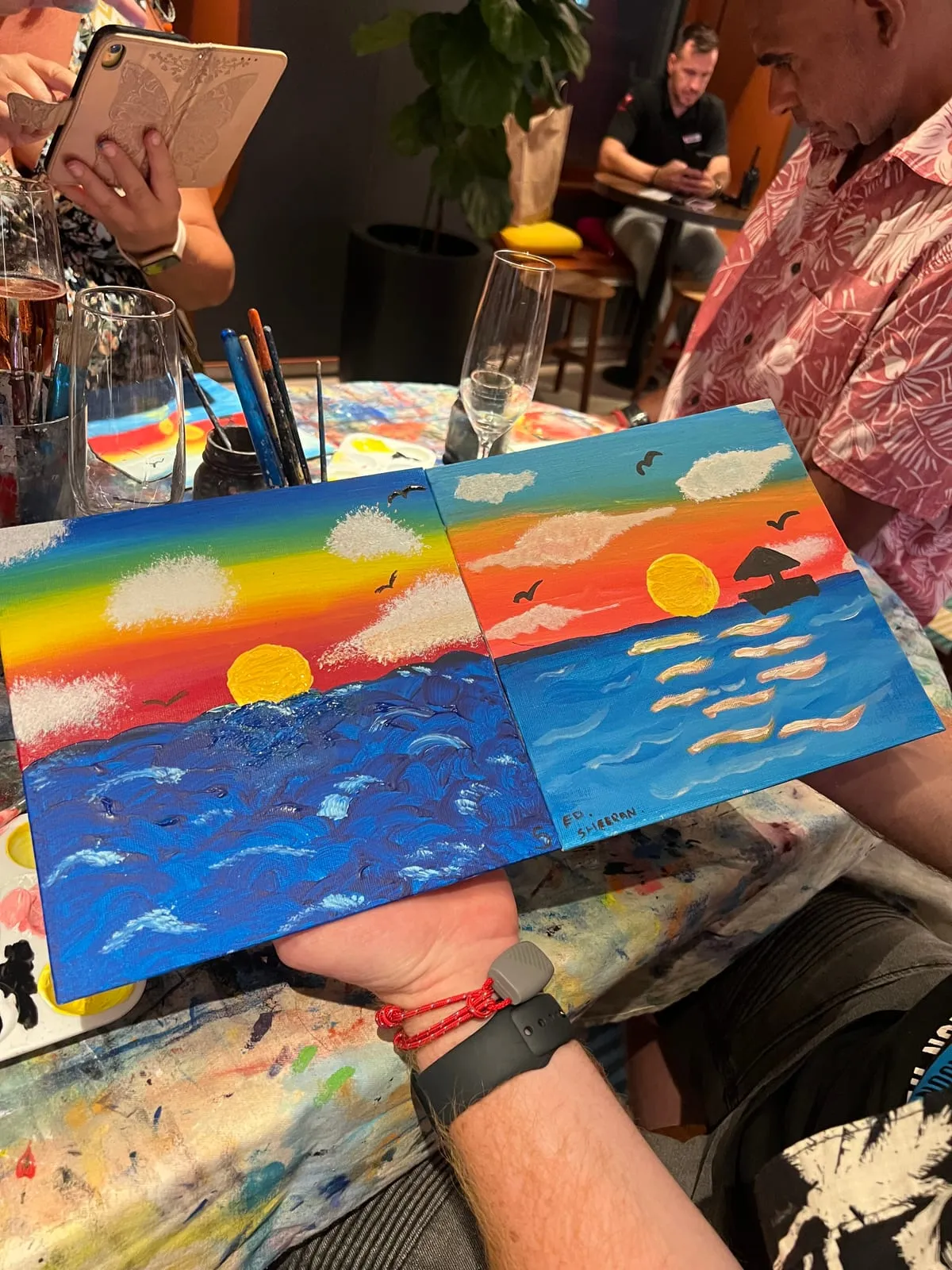 Our two finished canvases held side by side, one a sunset with a sailboat and a signature in the corner, the other a rainbow sky over a thickly painted sea