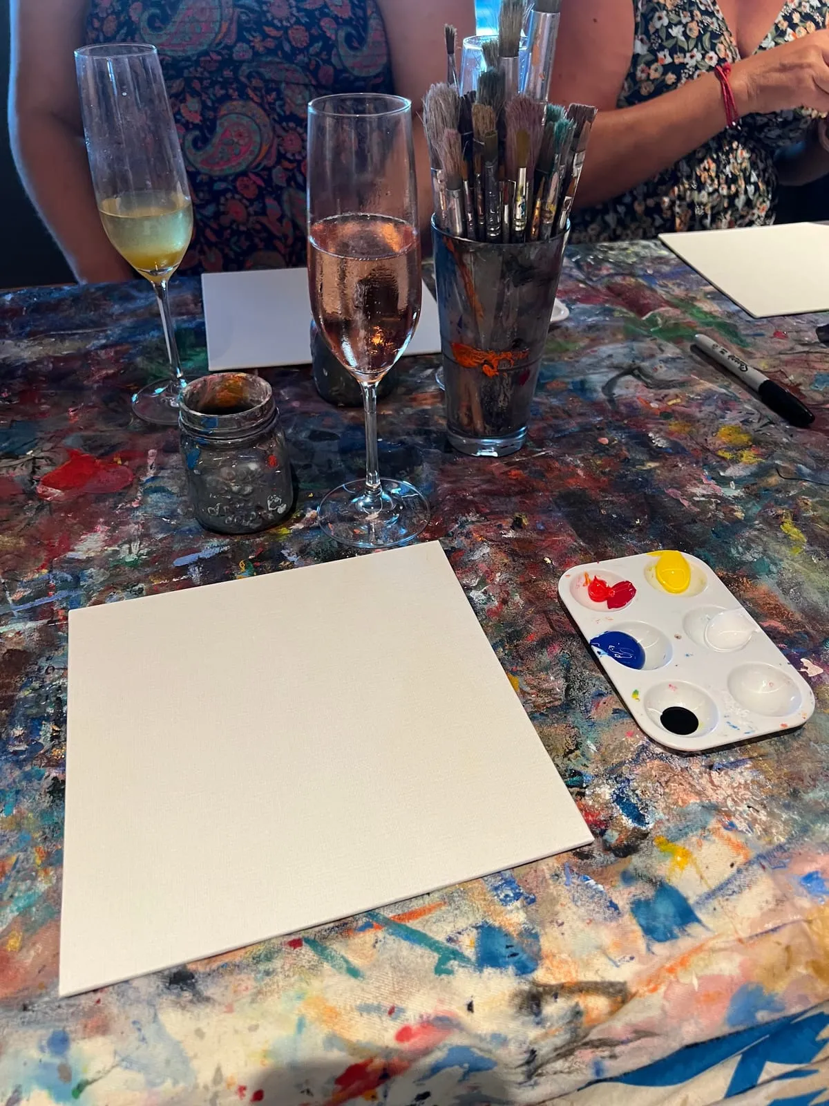 A blank canvas on a paint-caked table next to a palette of red, yellow, blue, black and white, a jar of brushes and two glasses of wine