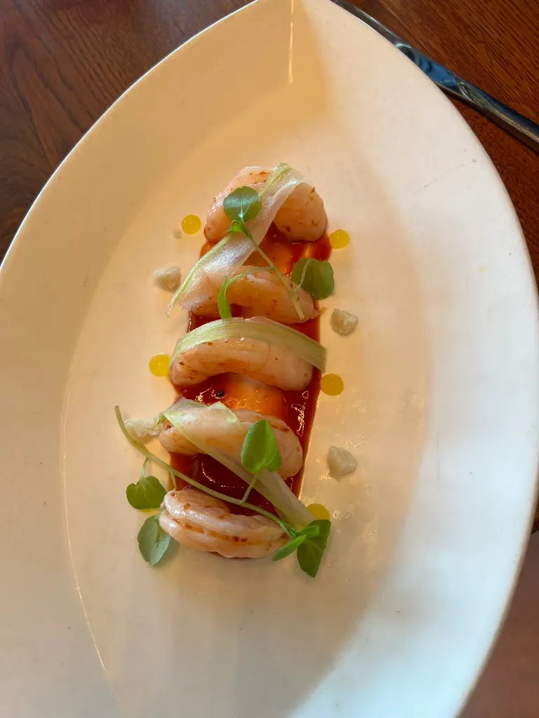 A prawn starter at The Wake: four prawns arranged along a smear of red sauce on a white oval plate, with shaved celery ribbons, micro leaves and dots of oil.
