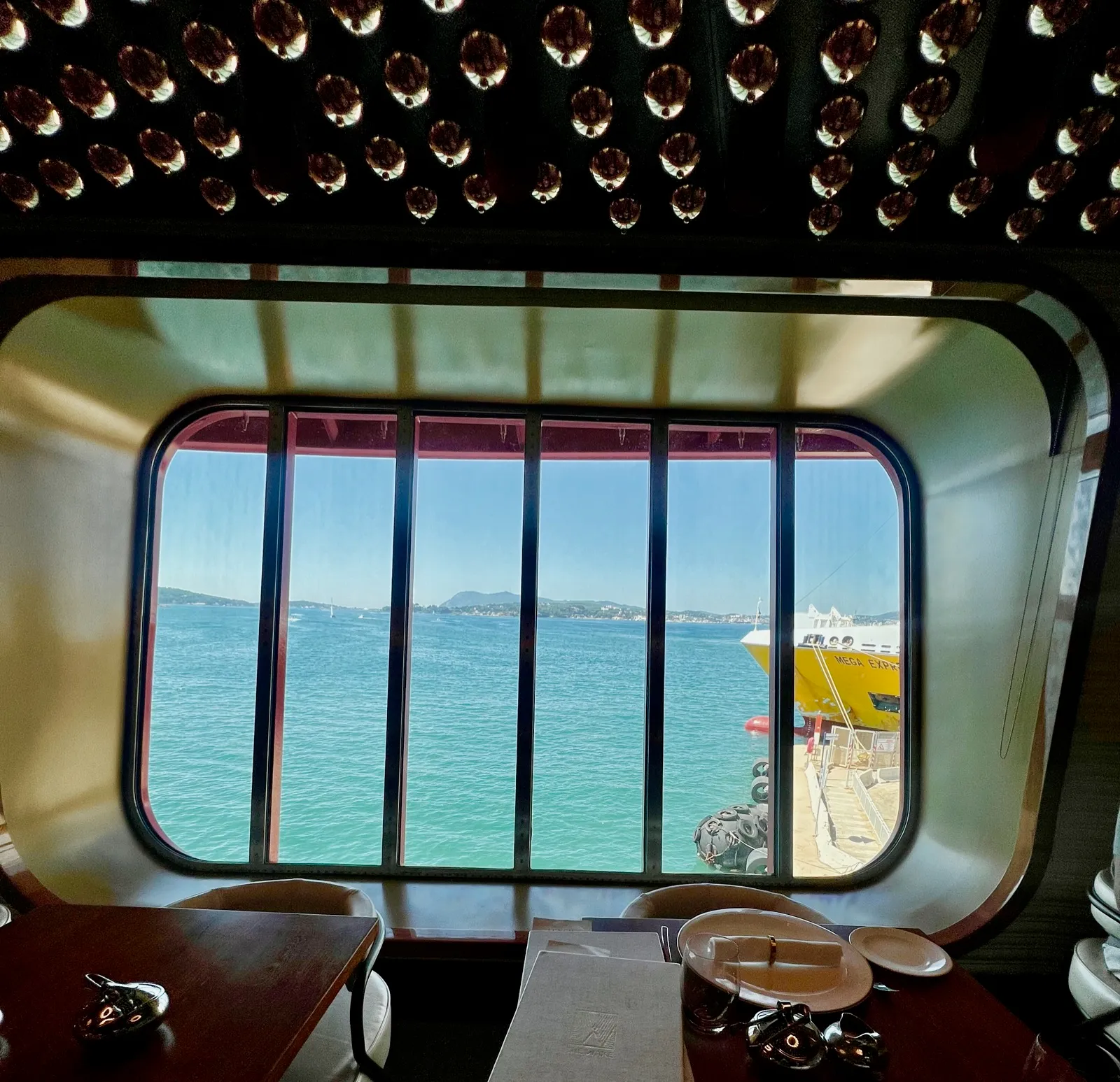 The view from a table at The Wake: a wide window looking out over turquoise sea to hills across the bay, the yellow Mega Express ferry tied up at the quayside, wine bottles racked in the ceiling above and the restaurant tables in the foreground.