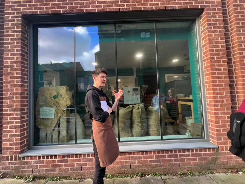 Our guide in a leather apron talking to the group on the pavement outside the roastery window, sacks of cocoa beans and the roasting machine visible through the glass behind him