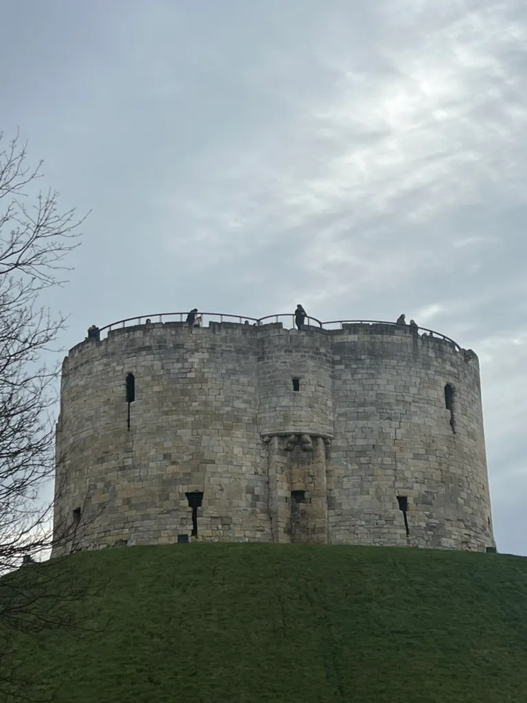 Clifford's Tower on its grassy mound, a squat round stone keep with people standing on the roof deck against a grey January sky