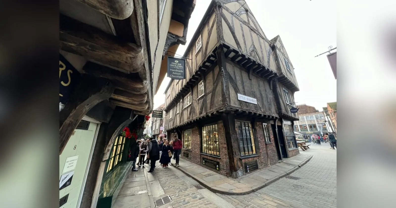 The corner of Little Shambles in York, leaning timber-framed buildings with a hanging chocolate shop sign and people walking down the narrow cobbled street
