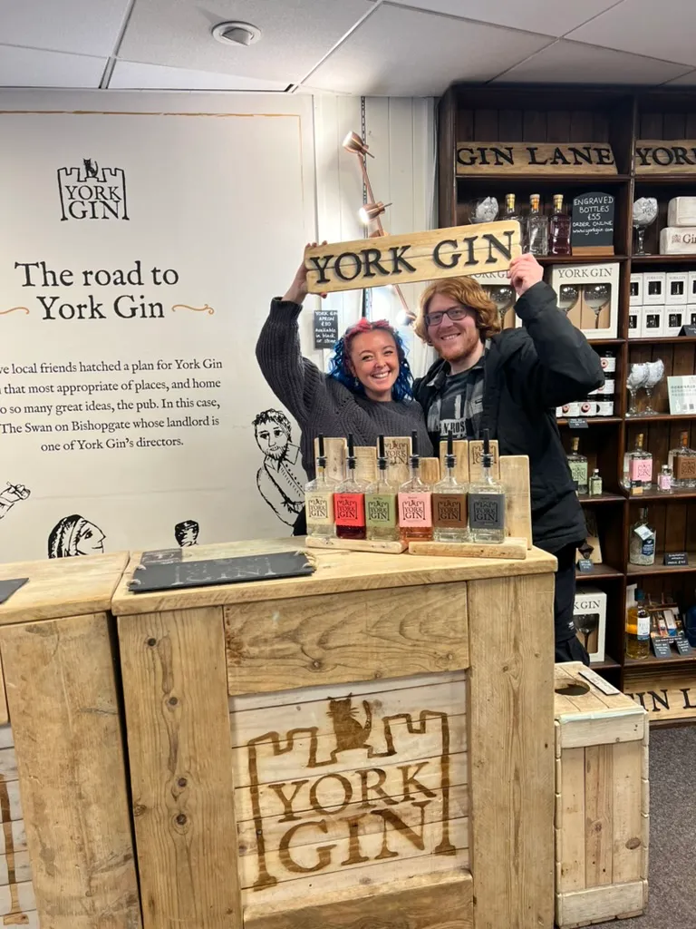 The two of us holding a burnt wooden York Gin sign above our heads in the shop, the six tasting bottles on the counter in front of us