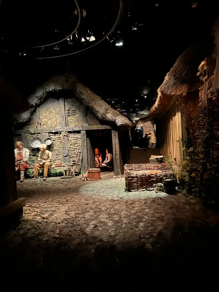 The reconstructed Coppergate street from the ride, thatched timber houses with two figures sitting outside one of them and a wicker fence in the foreground