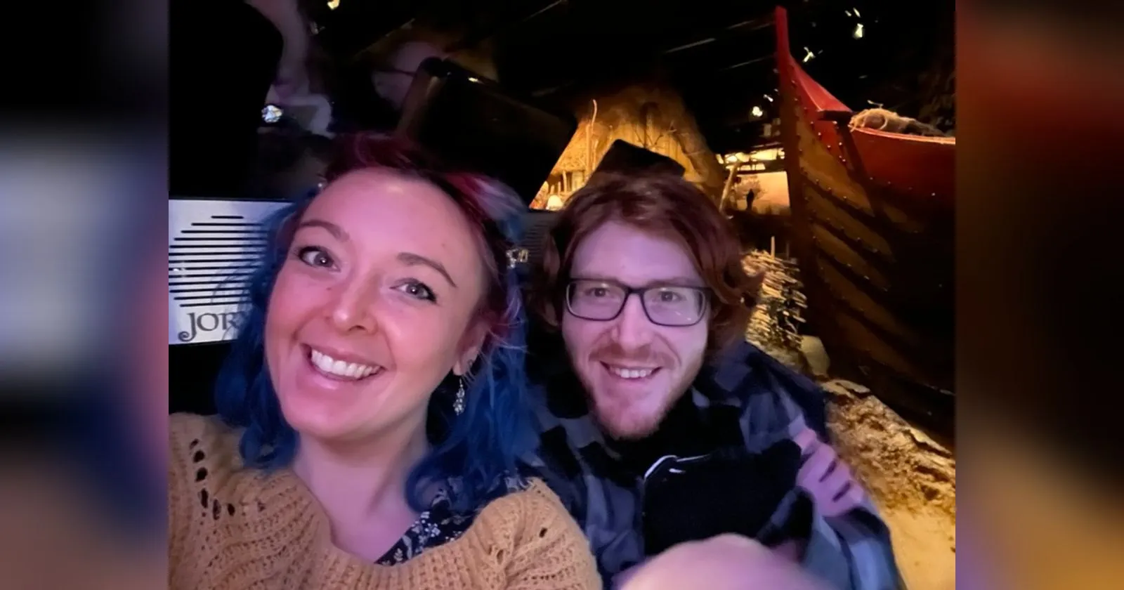 The two of us grinning in a JORVIK time capsule with a red-painted Viking boat and a thatched roof lit up behind us