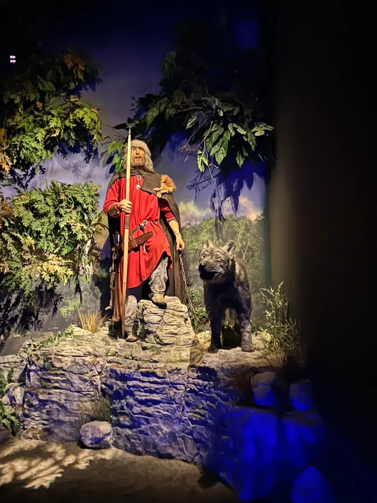 A Viking figure in a red tunic holding a spear on a rocky outcrop with a grey wolf on a chain beside him, lit blue and green