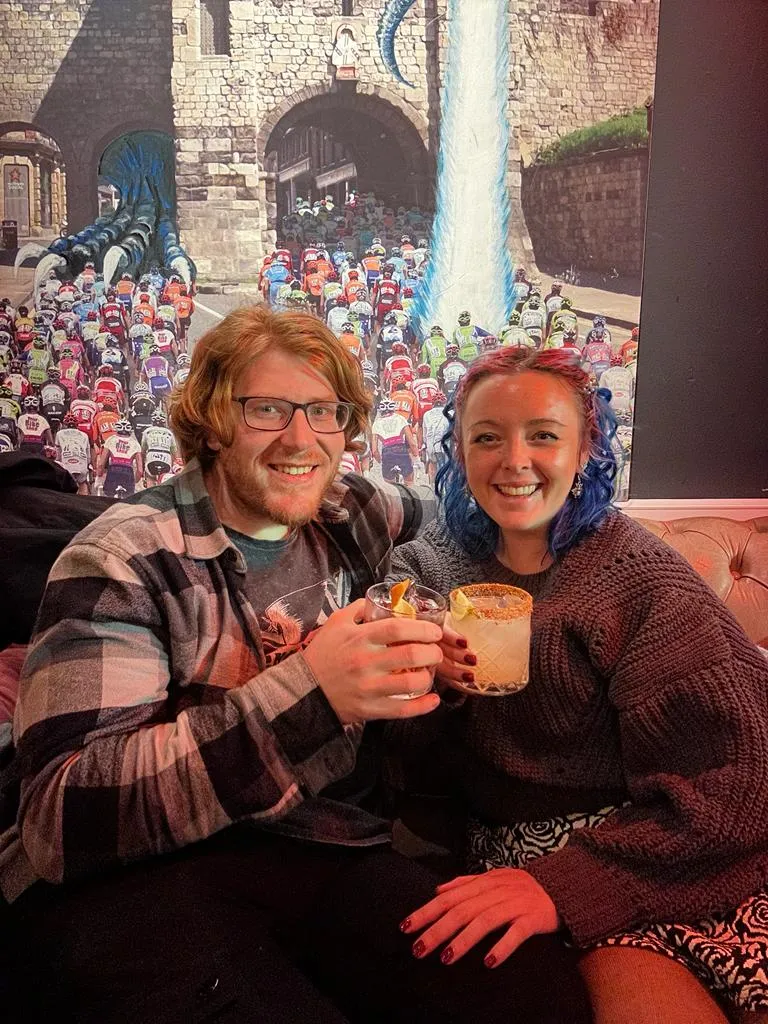 The two of us on a leather sofa with two cocktails, a large painting of a peloton of cyclists riding through a stone city gateway on the wall behind