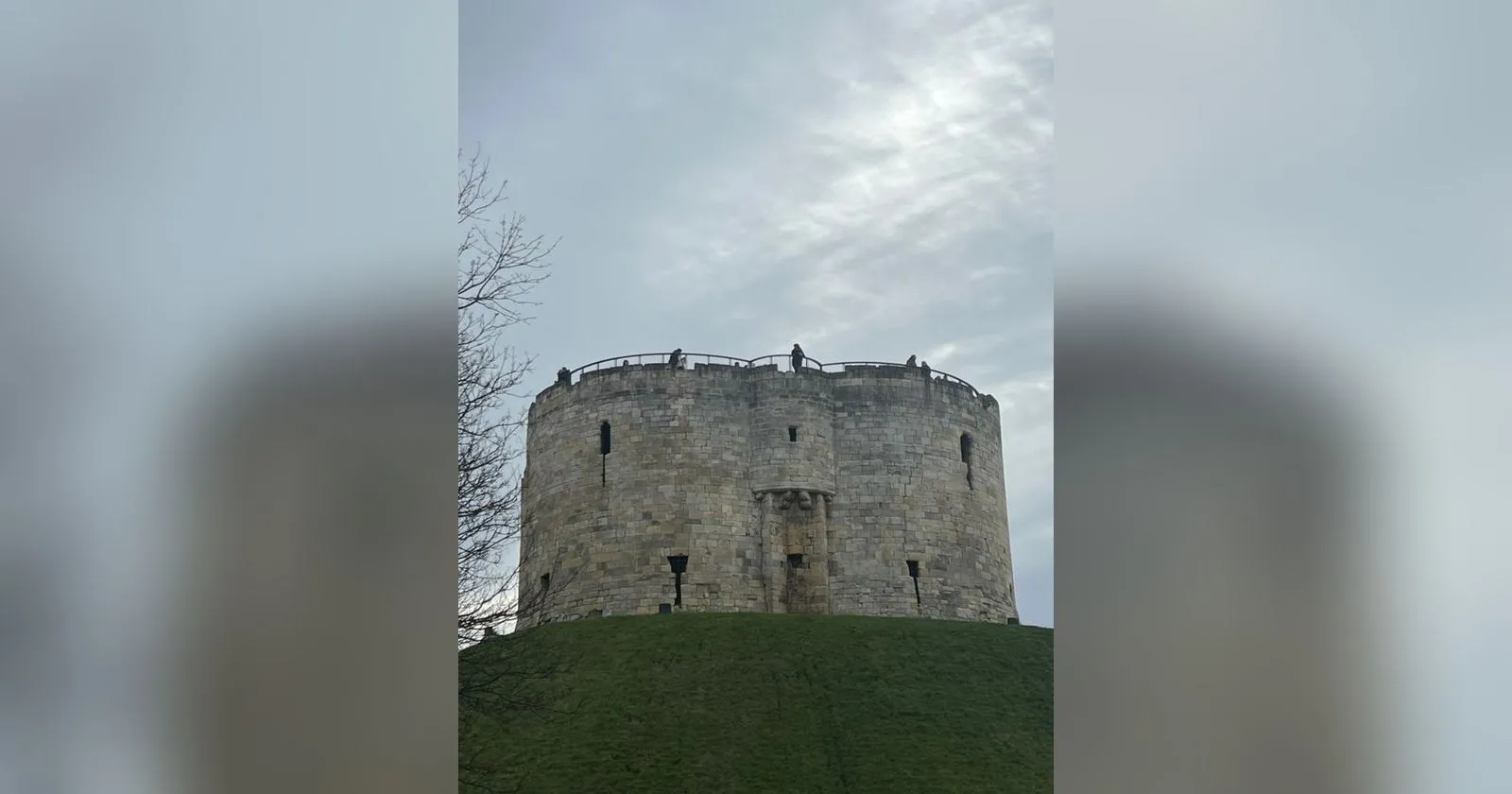 Clifford's Tower on its grassy mound, a squat round stone keep with people on the roof deck against a grey January sky