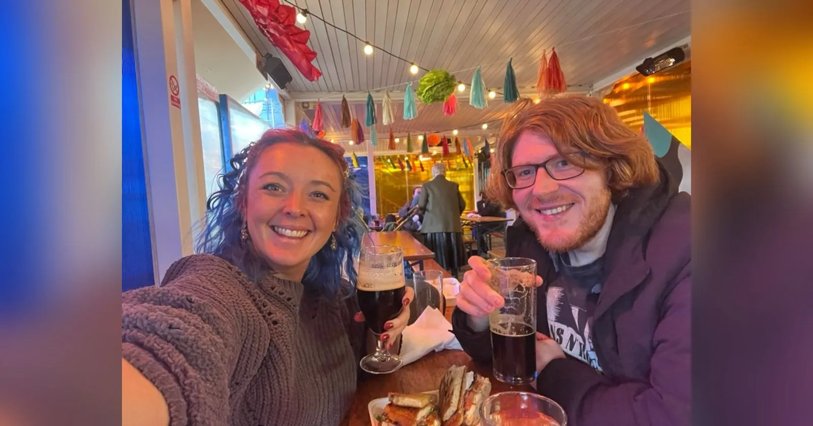 The two of us at a wooden table under paper tassels and festoon lights at Spark:York, pints raised and a basket of chicken sandwiches on the table