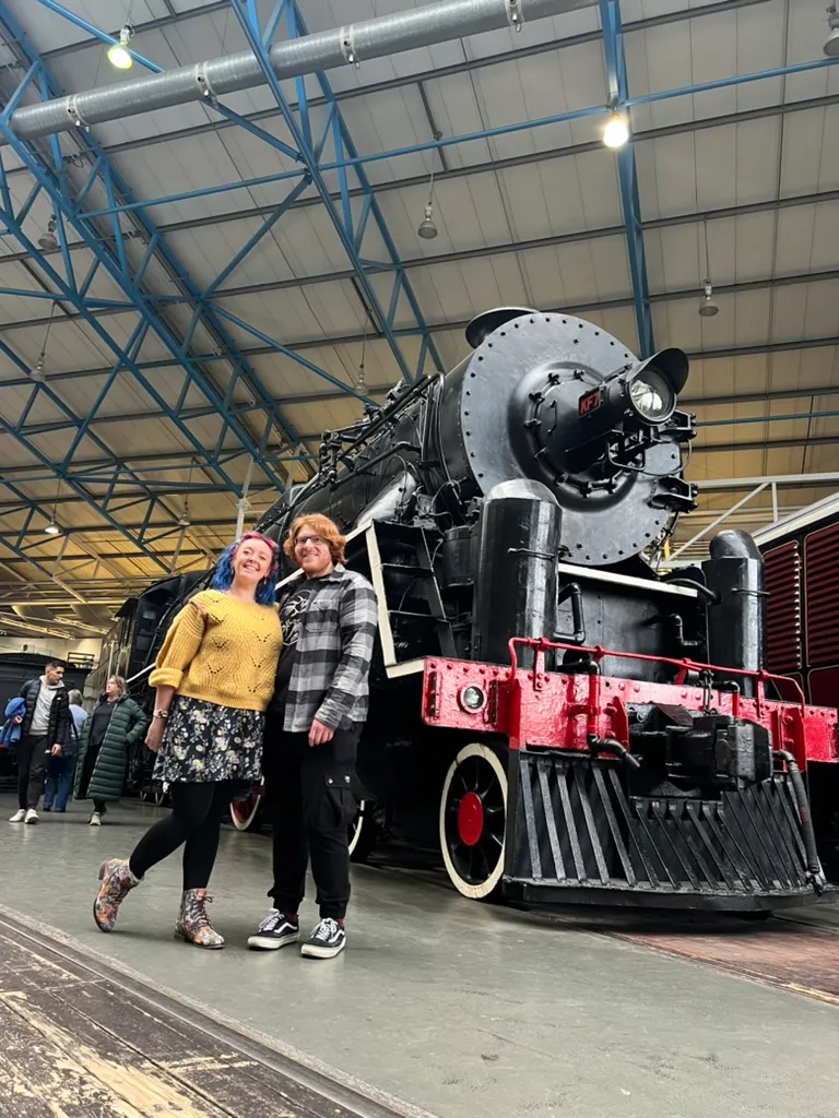 The two of us standing beside an enormous black steam locomotive with red trim in the Great Hall, the blue roof girders overhead