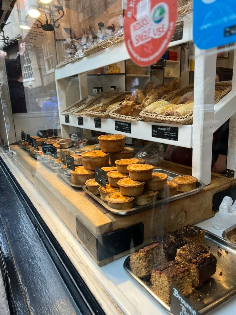 A bakery window in the Shambles packed with rows of golden pork pies on wooden boards, baskets of pasties and sausage rolls above them and a tray of brownies below