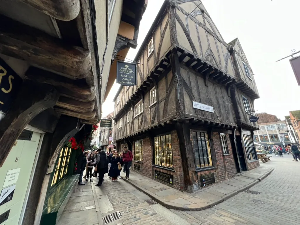 The corner of Little Shambles, leaning timber-framed buildings with a hanging sign for a chocolate shop and people walking down the narrow cobbled street