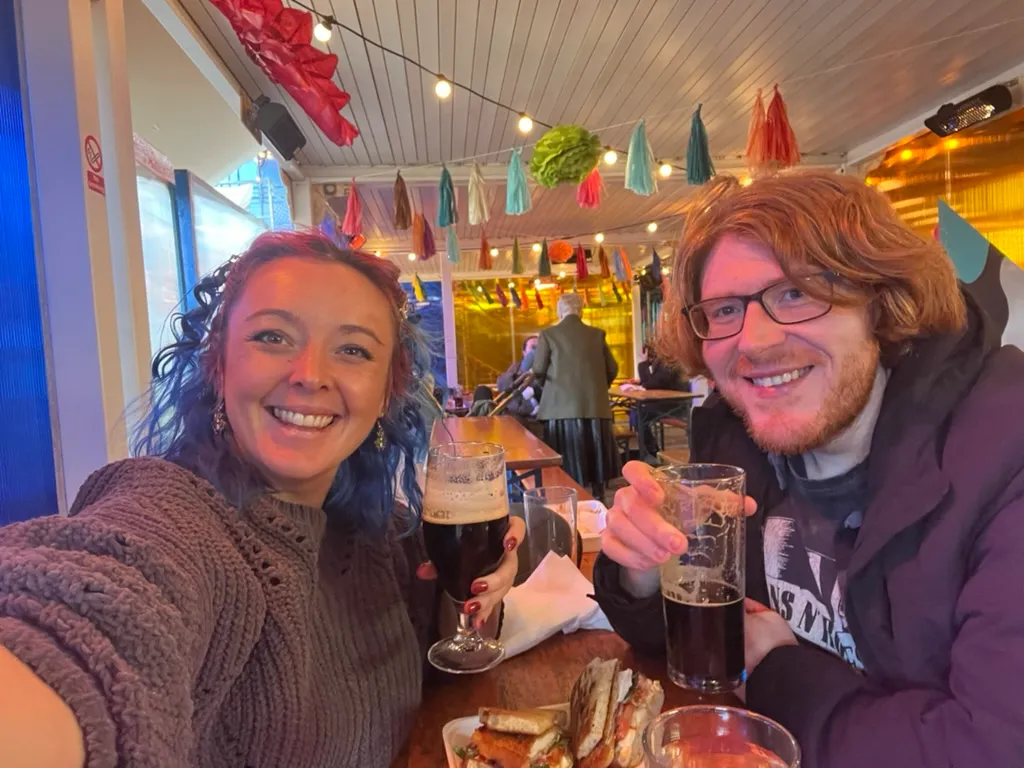 The two of us at a wooden table under strings of colourful paper tassels and festoon lights at Spark:York, a dark stout and a pint of ale raised between us and a basket of toasted chicken sandwiches on the table