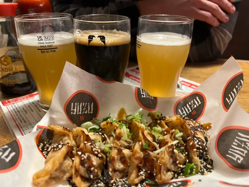 A tray of gyoza covered in sesame seeds, spring onion and sauce, with three glasses of beer lined up behind it, two pale and one dark